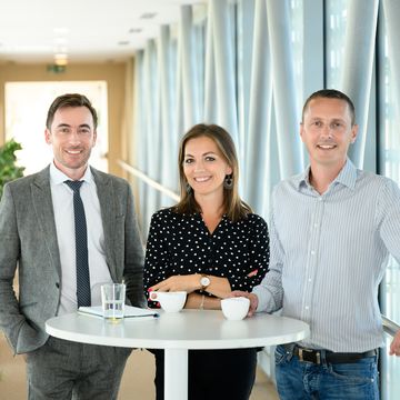 Három kolléga áll egy asztalnál kávézás közben és beszélgetnek.